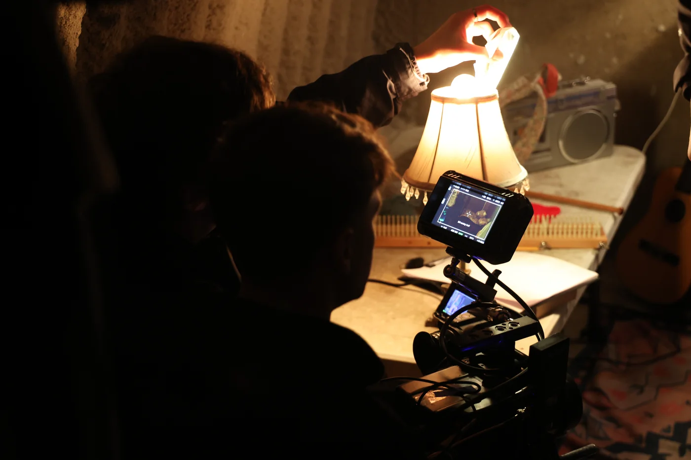 Crew silhouetted around a camera and monitor pointed at a lamp-lit desk covered in objects