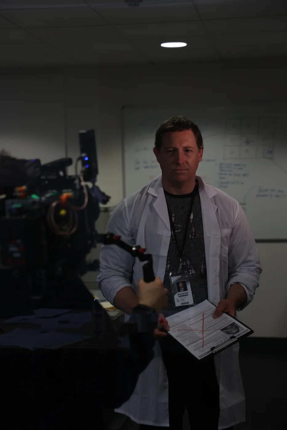 An actor in a white lab coat with an ID lanyard holds a clipboard in a strip-lit room, a film camera in the foreground